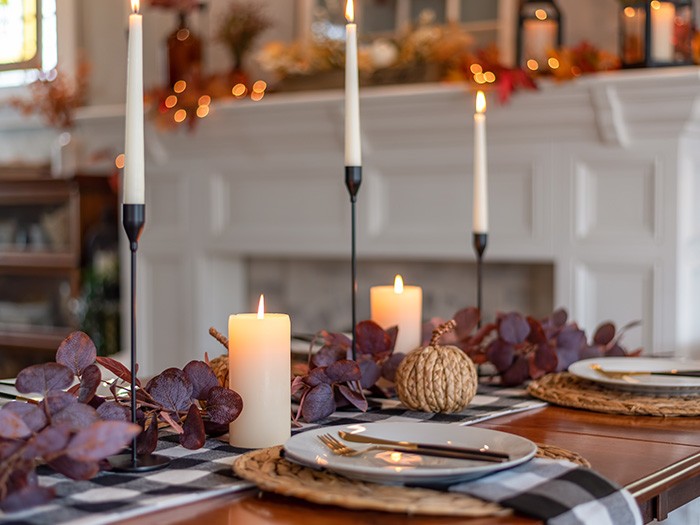 A fall-themed tablescape featuring dried eucalyptus leaves in rich red hues, modern black candle holders, and rustic roped pumpkin decor. The setting overlooks a white fireplace adorned with additional autumn leaf accents, creating a warm and inviting atmosphere.